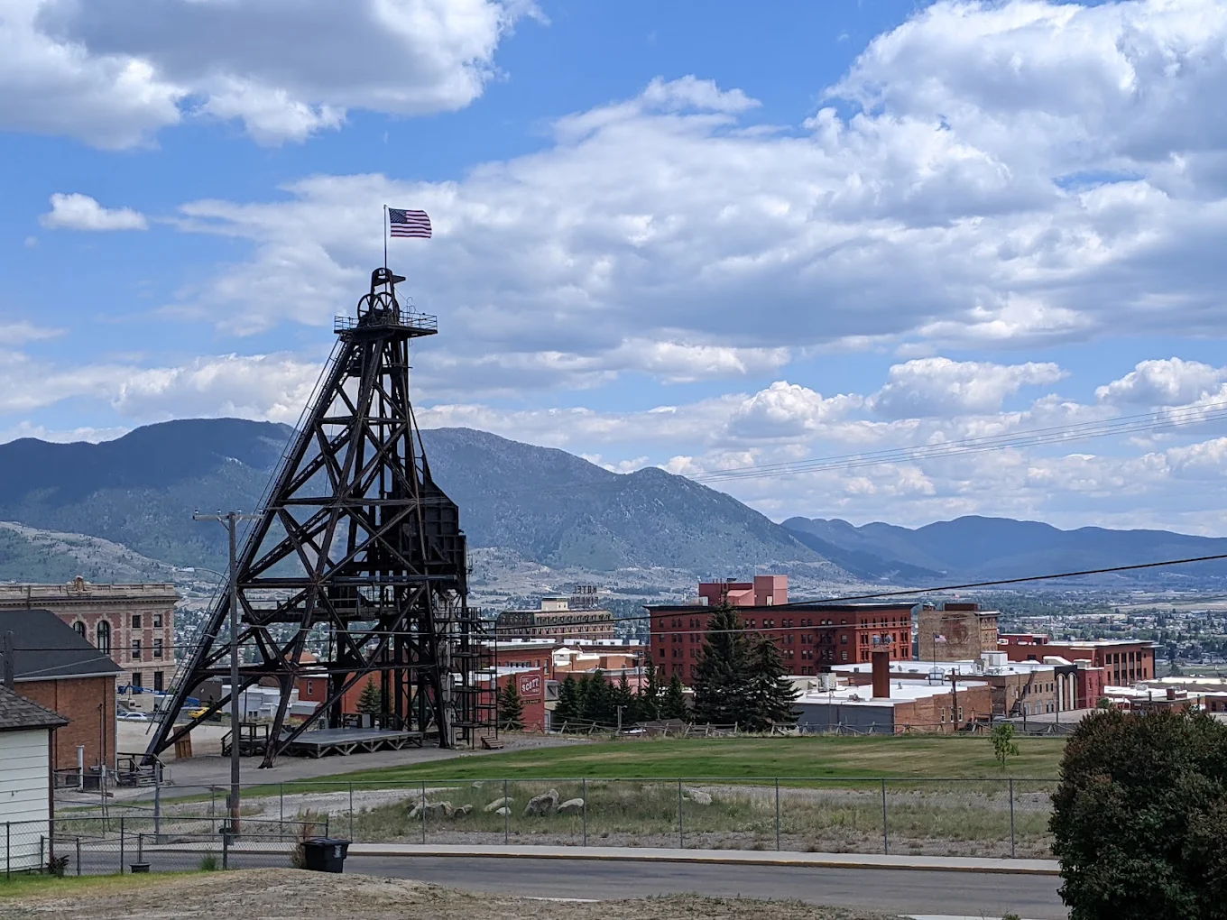 A mining headframe with an American flag stands near red brick buildings, with mountains and a cloudy blue sky in the background.