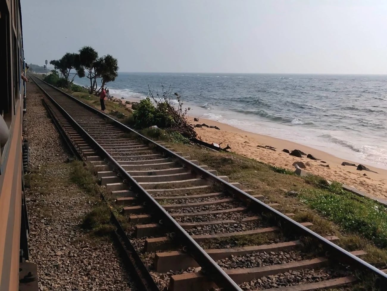 View from a train window of railway tracks running parallel to a sandy beach with ocean waves, green bushes, and a few trees under a bright sky.