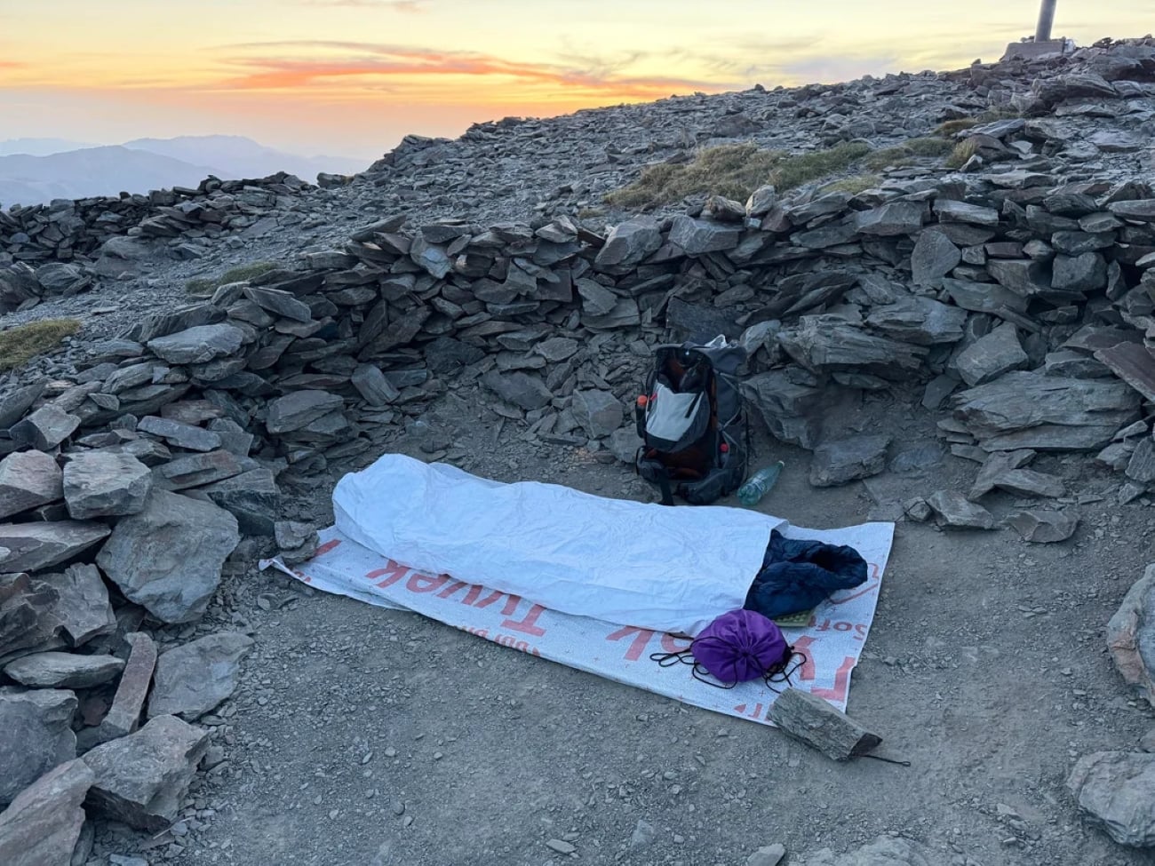 Photos posted by hikers often capture moments like this: a sleeping bag on a mat inside a rocky shelter at sunrise, with a backpack and gear nearby, set against a stunning mountain landscape.