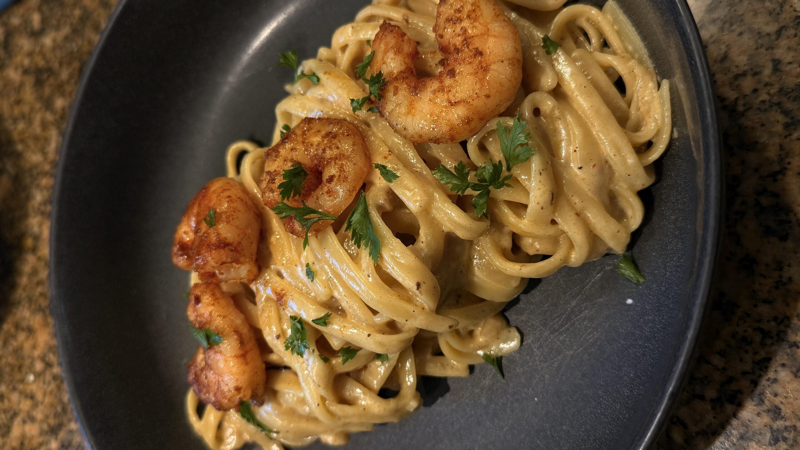 Fettuccine pasta in a creamy sauce topped with seasoned shrimp and garnished with fresh parsley, served in a dark bowl on a granite countertop—a dish as delicious as it is photogenic, perfect for those who love sharing photos of pasta.