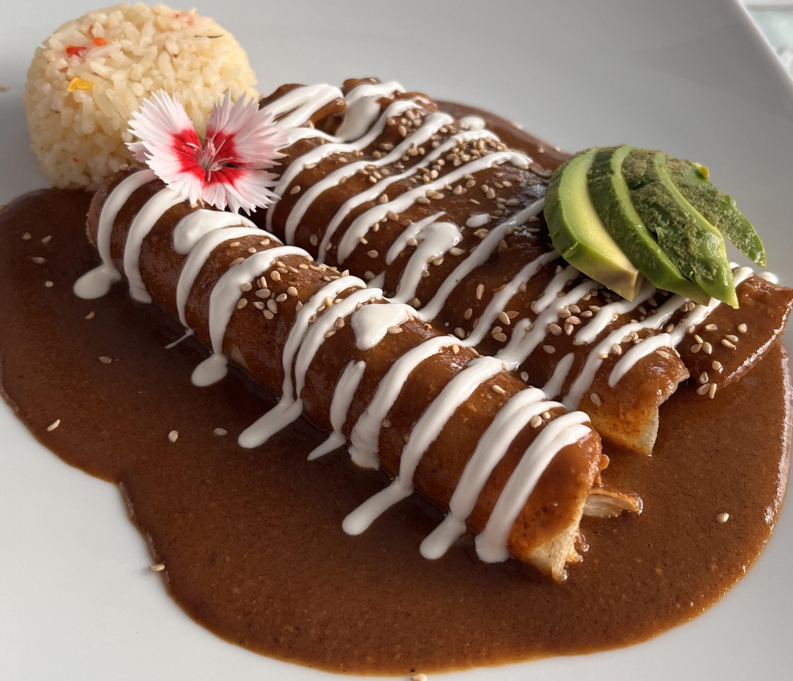 Two enchiladas covered in mole sauce and drizzled with cream, topped with sesame seeds and avocado slices, served with a flower garnish and a round portion of rice on a white plate.