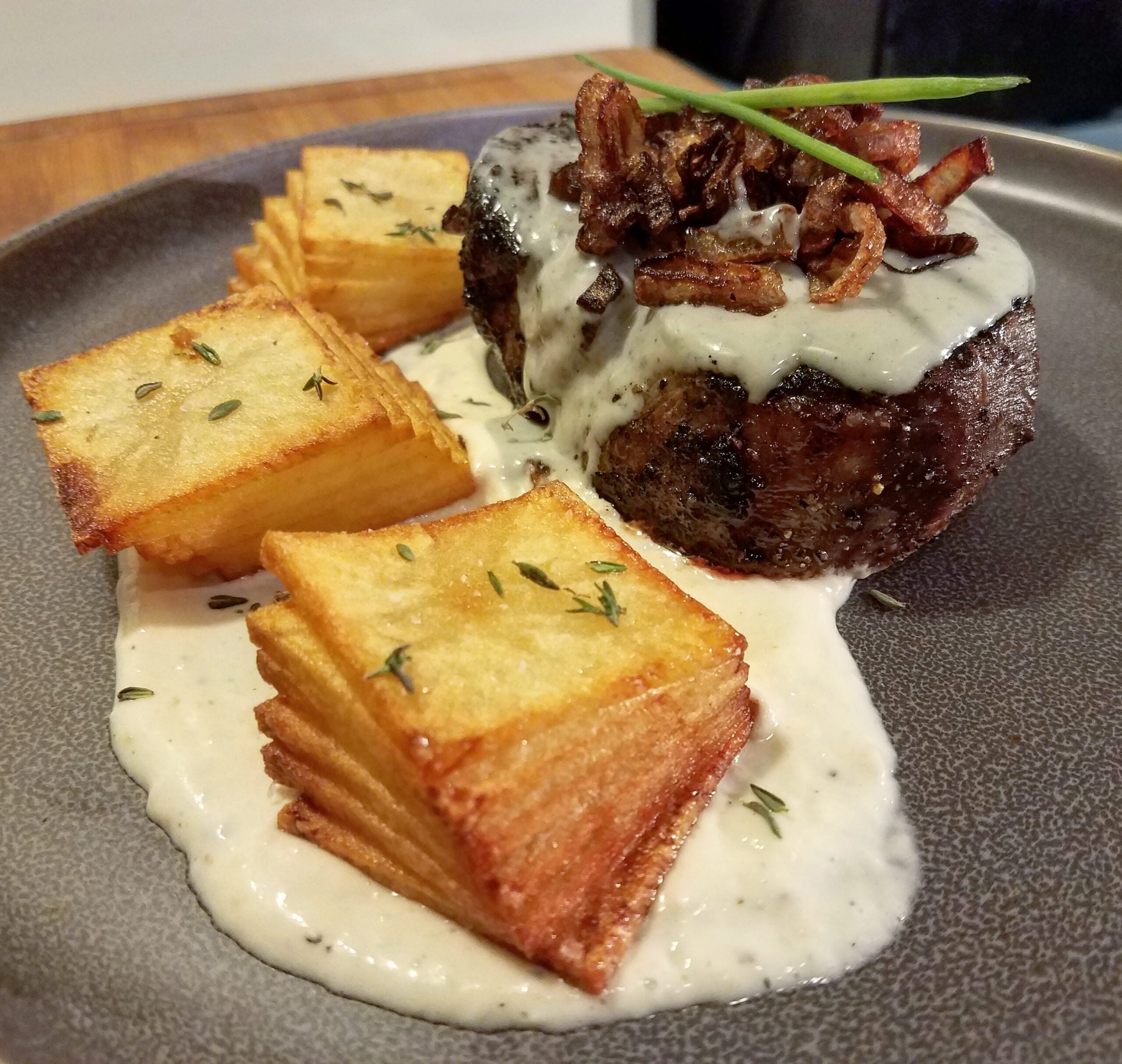 A plated dish featuring stacked crispy potato cubes, a steak topped with creamy sauce and fried onions, garnished with chives and thyme, served on a gray plate.