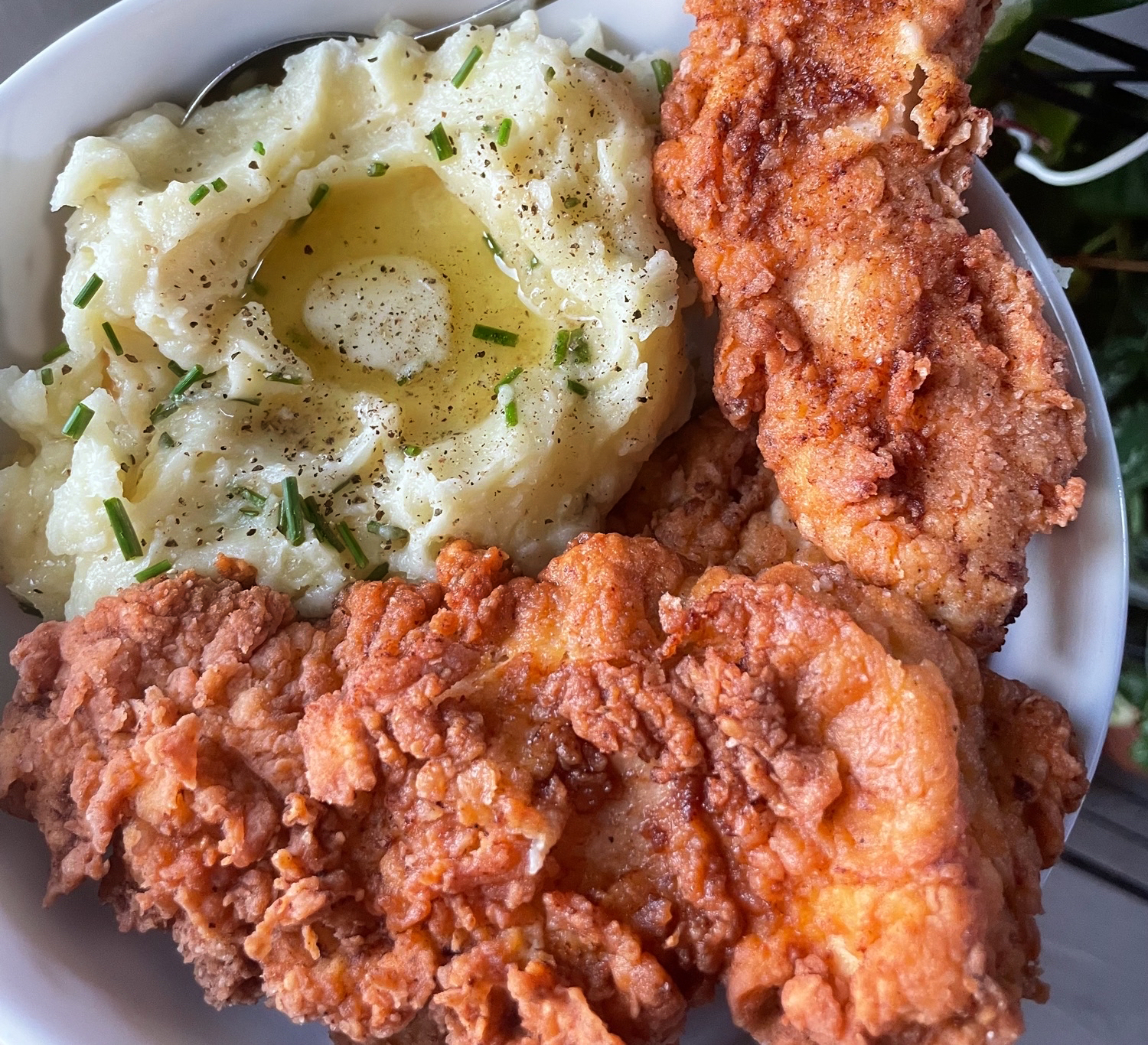 A bowl of creamy mashed potatoes topped with butter, pepper, and chives, served alongside two pieces of crispy fried chicken.