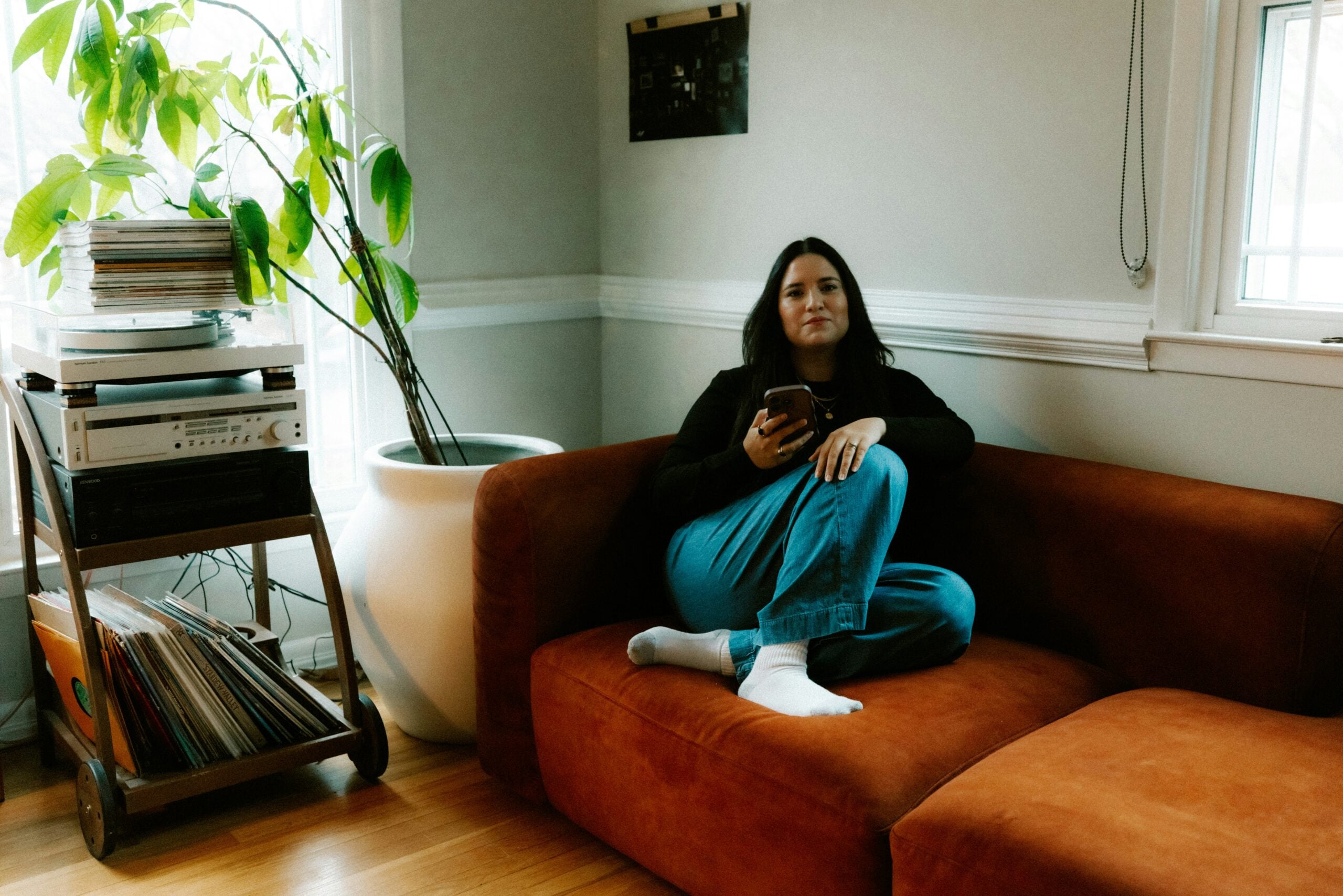 A person sits casually on a rust-colored couch, holding a phone. Nearby is a stand with a stereo and records, and there’s a large potted plant by the window. The room has light walls and wood floors.