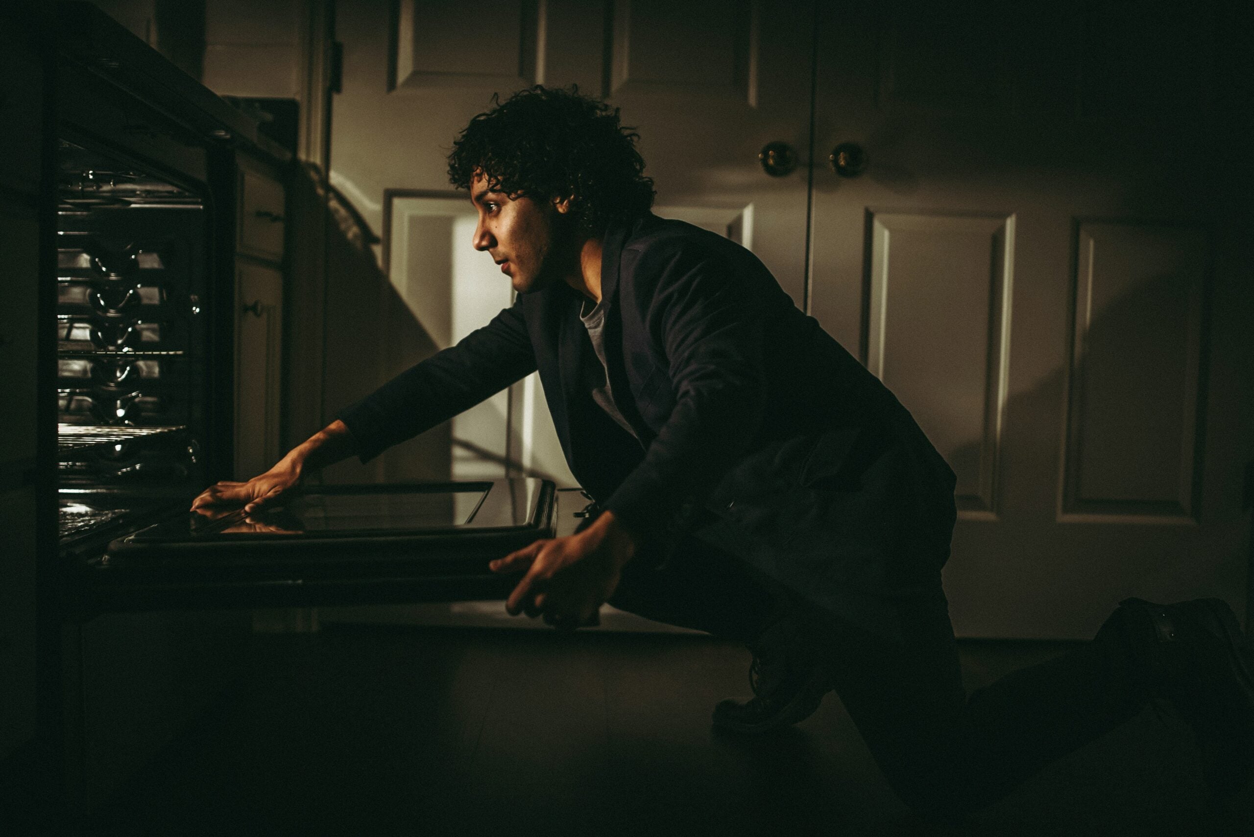 A person kneels in front of an open oven, reaching inside with one hand. The scene is dimly lit, creating a dramatic and focused atmosphere in a kitchen.