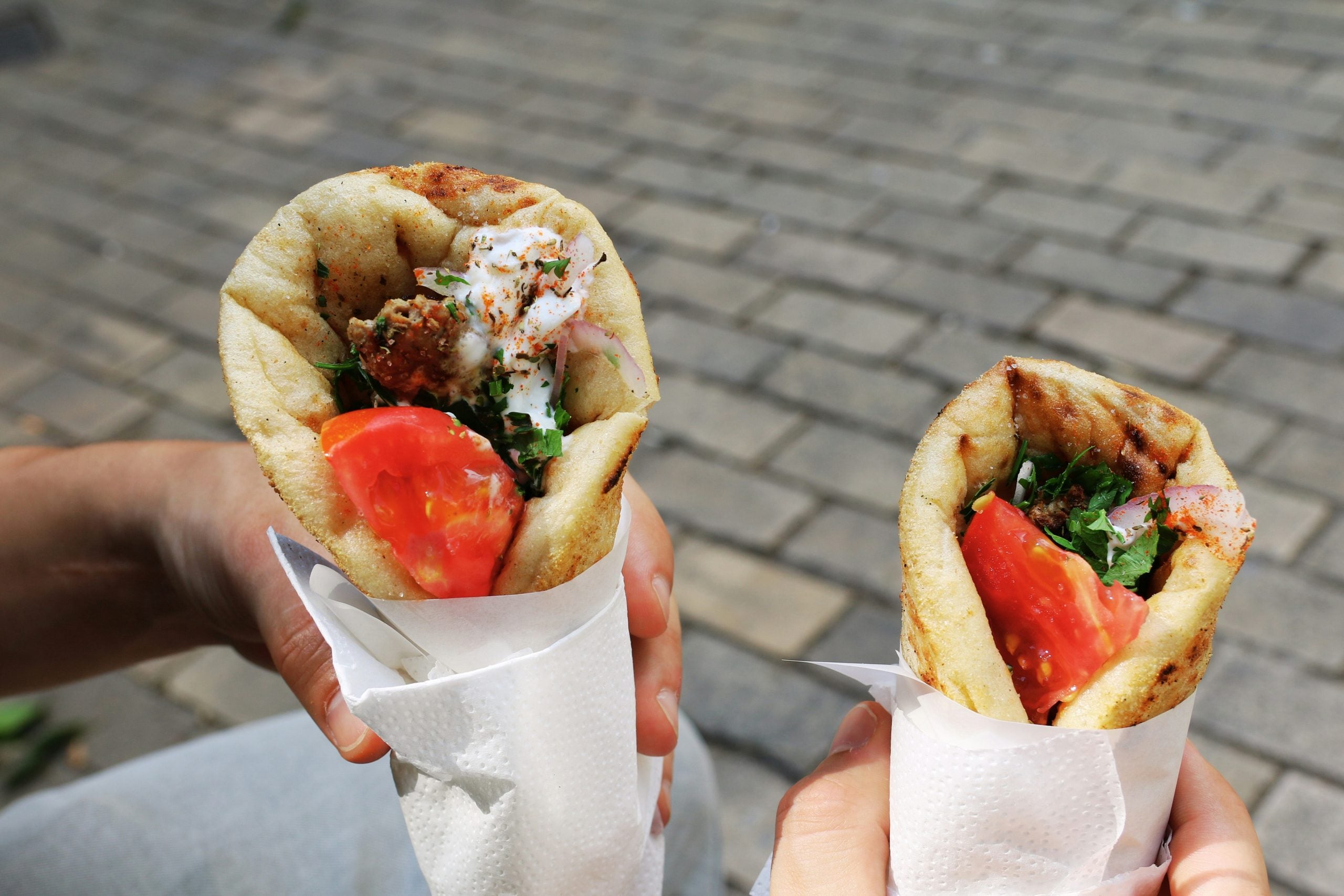 Two hands holding wrapped pita sandwiches filled with grilled meat, fresh tomato slices, onions, and herbs, against a blurred outdoor brick pavement background.