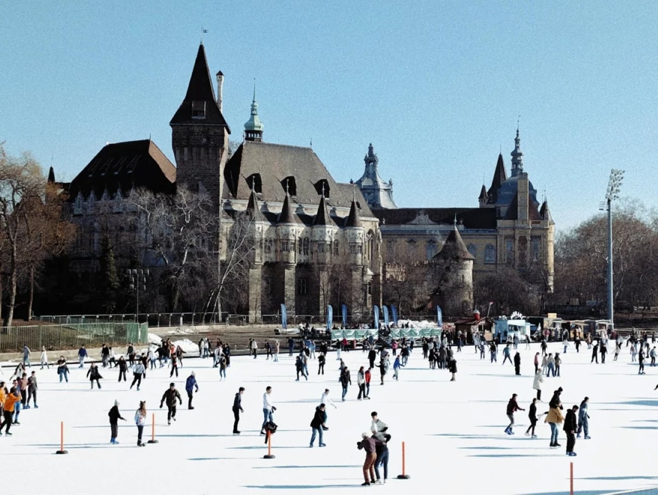People ice skate on a large outdoor rink in front of a historic castle-like building with towers on a clear day. Trees without leaves frame the scene.