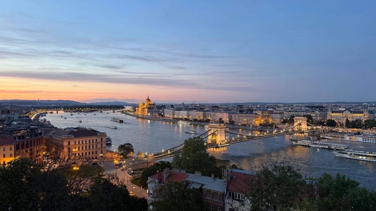 A panoramic view of Budapest at sunset, featuring the Chain Bridge over the Danube River and the illuminated Hungarian Parliament building in the distance.