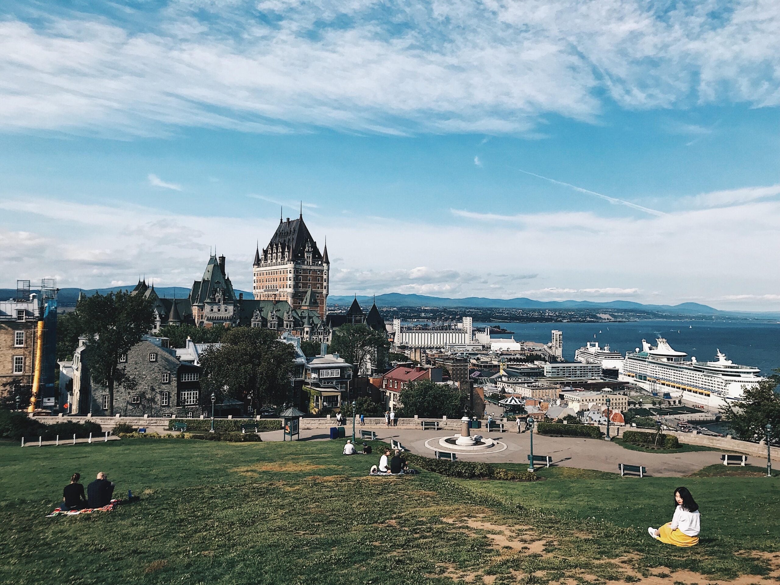 A panoramic view of Quebec City showing Château Frontenac, cruise ships docked at the port, and people relaxing on a grassy hillside under a blue sky with scattered clouds.