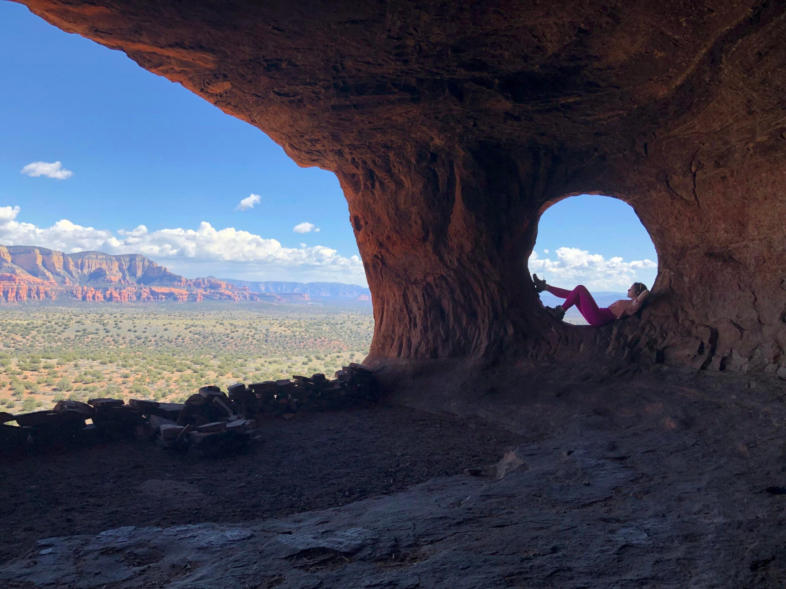 A person lounges inside a circular window of a rock cave, overlooking a vast desert landscape with red rock formations and a blue sky dotted with clouds—an iconic spot where hikers often pause to take photos posted online, capturing the adventurous spirit of the scene.