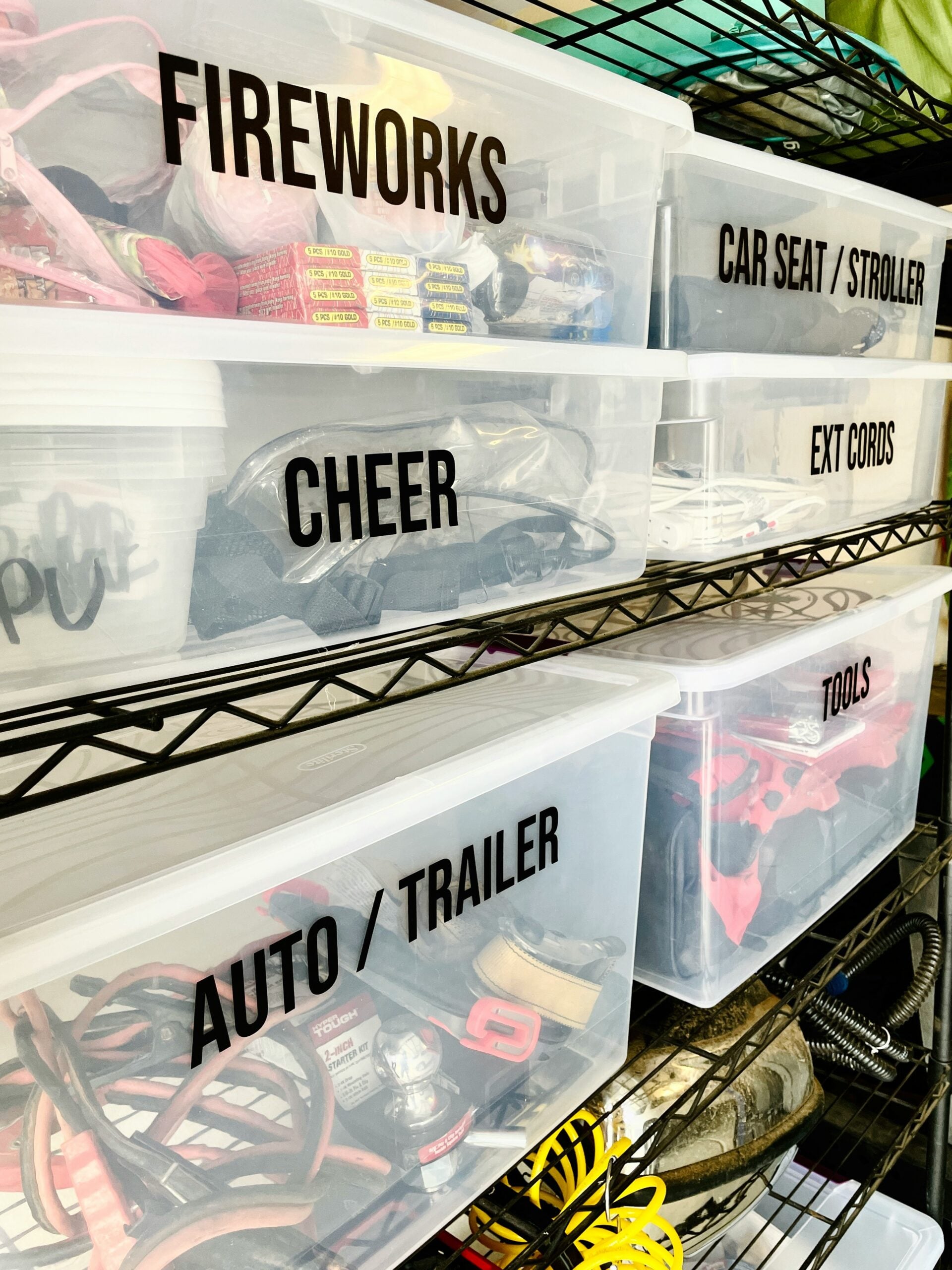 Plastic storage bins on metal shelves, each labeled for contents: FIREWORKS, CHEER, AUTO/TRAILER, CAR SEAT/STROLLER, EXT CORDS, and TOOLS. The bins hold various items related to the labels.