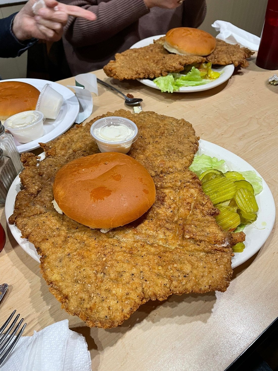 A large breaded pork tenderloin sandwich with pickles and lettuce on a plate, dwarfing the bun. Another similar plate and hands are visible in the background.