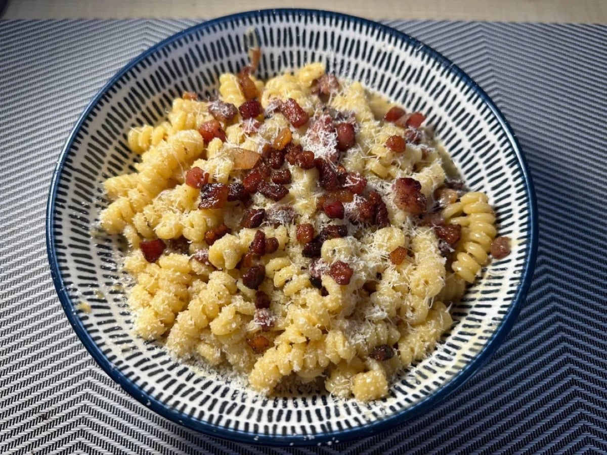 A bowl of fusilli pasta topped with crispy bacon bits and grated cheese sits on a blue and white patterned plate on a striped placemat, capturing the delicious details seen in many inviting photos of pasta.