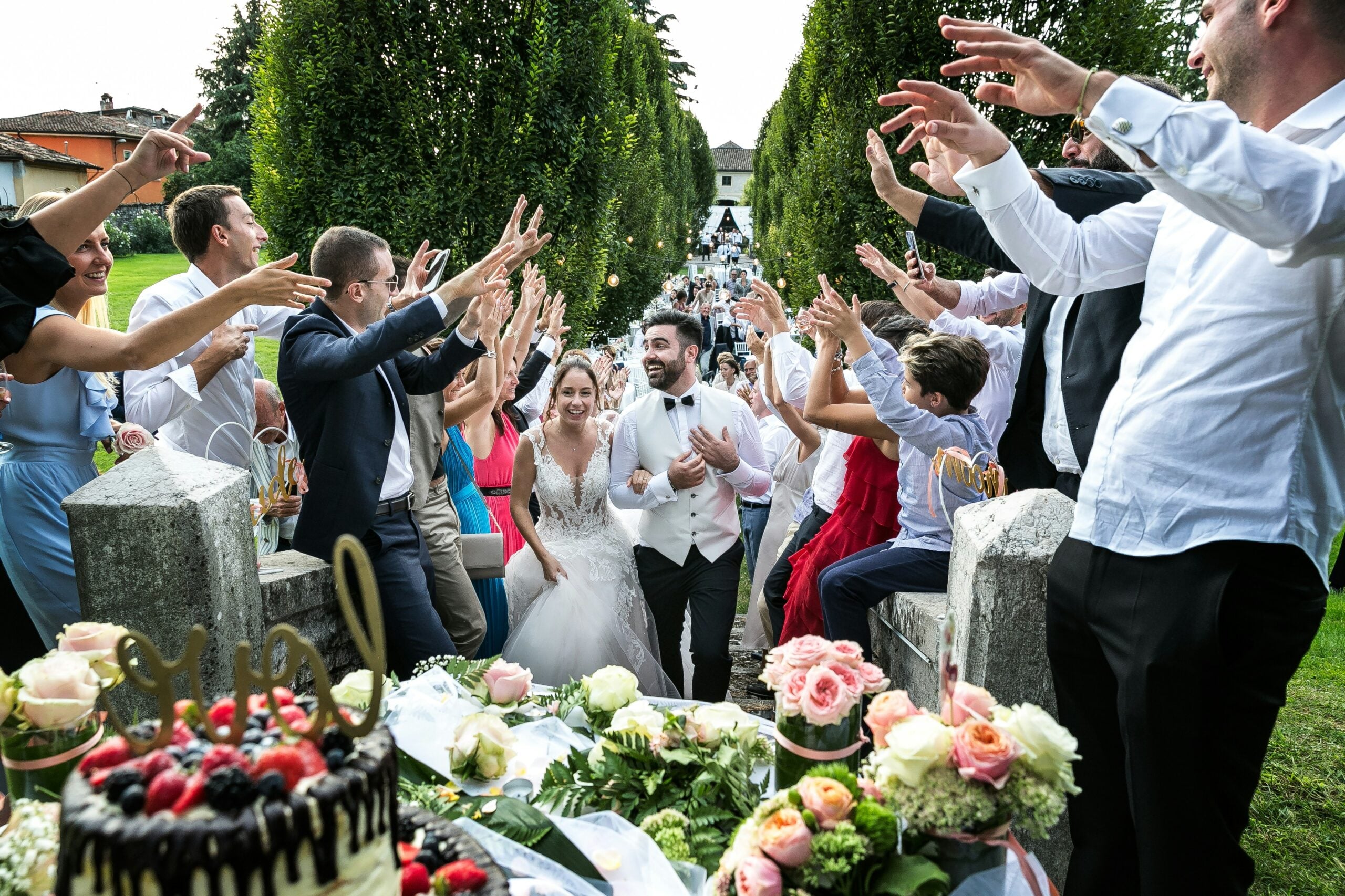 A bride and groom walk through a cheering crowd outdoors, surrounded by smiling guests with raised arms, flowers, and a cake in the foreground.