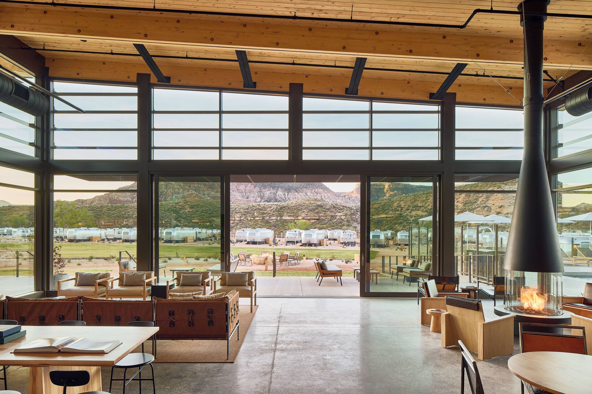 Spacious lodge with modern furniture, large glass windows showcasing a campground with trailers, surrounded by mountains. Offering the comforts typically found in top resorts, the indoor fireplace and cozy seating create an inviting atmosphere for relaxation.