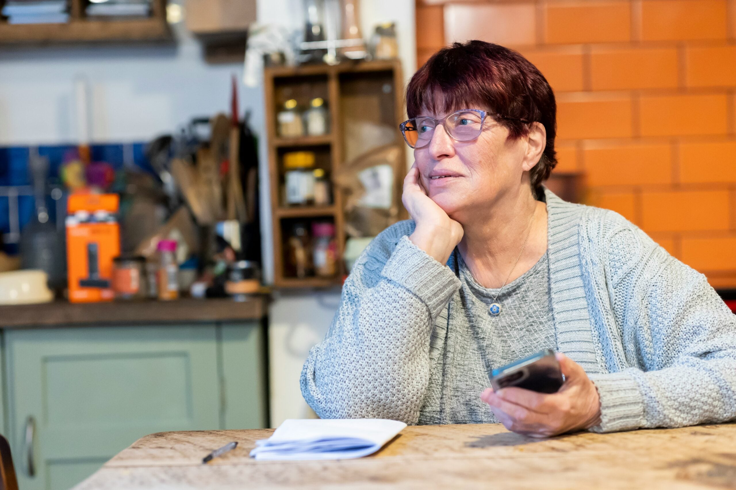 Older woman with short red hair and glasses sits at a kitchen table, holding a smartphone and notebook, looking thoughtfully to the side as she considers her retirement savings. Kitchen shelves and orange tiles are visible in the background.