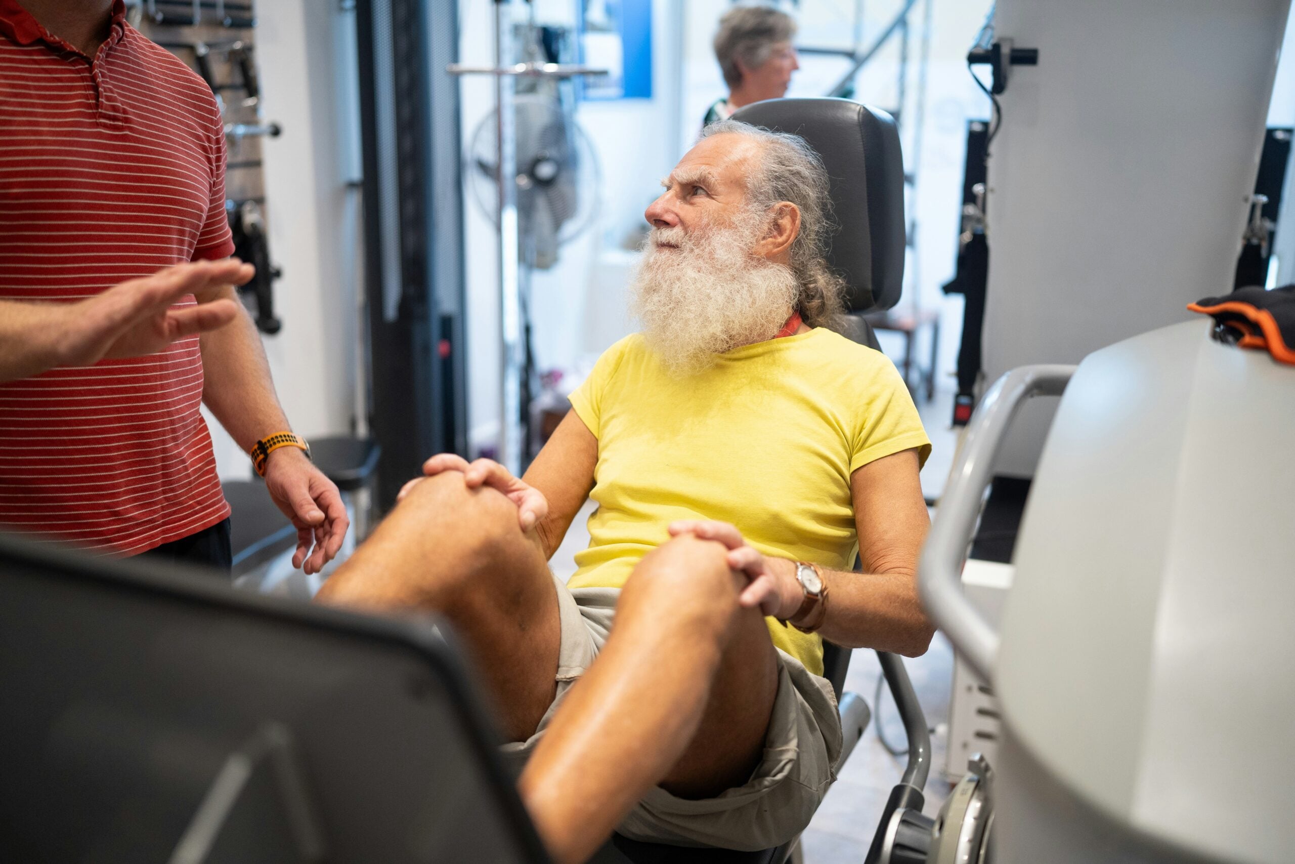 An older man with a white beard, wearing a yellow shirt and shorts, uses a leg press machine at a gym while discussing retirement savings with another person standing nearby.