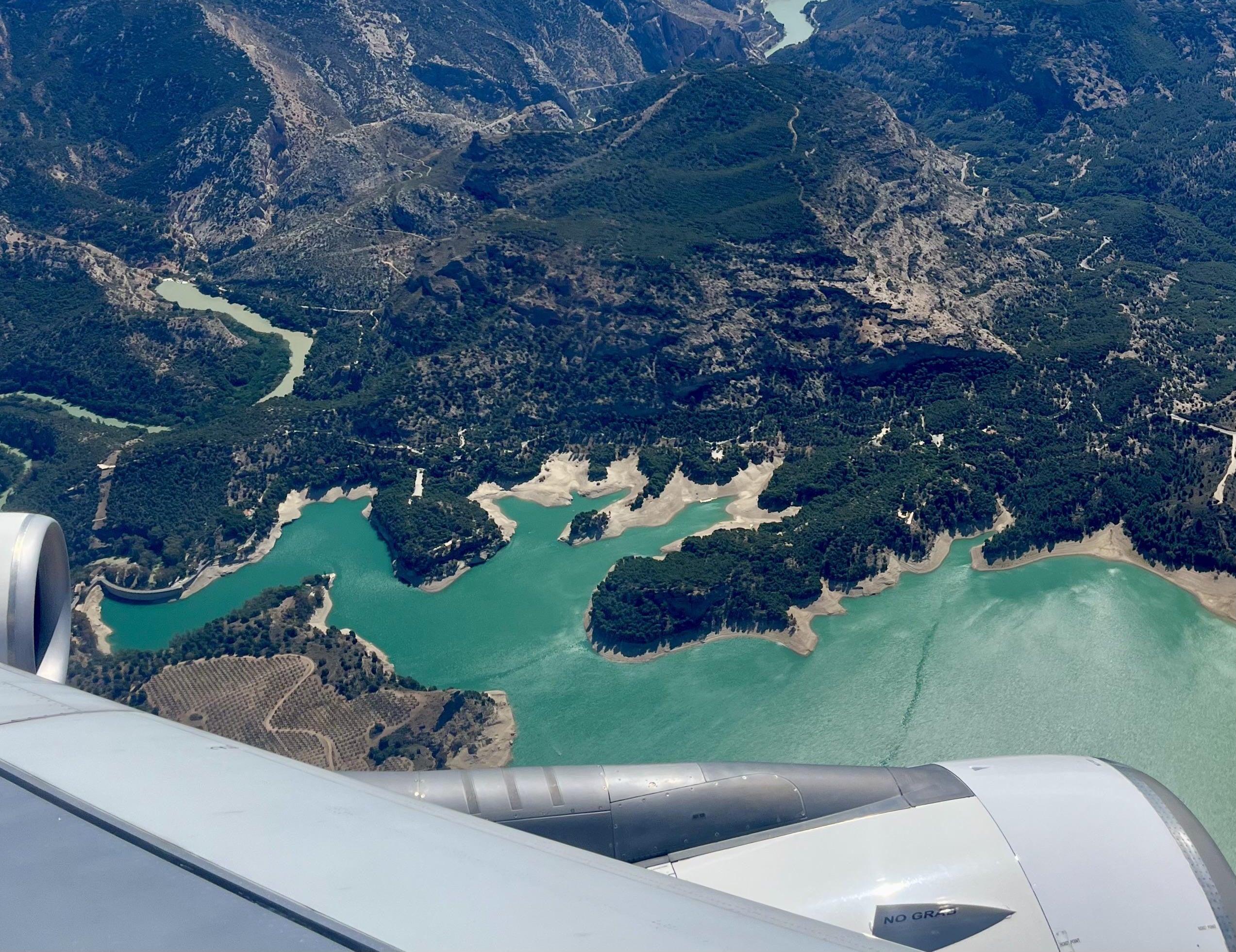 Aerial view from airplane windows showing a green lake surrounded by forested mountains and rugged terrain, with part of the aircraft wing visible in the foreground.