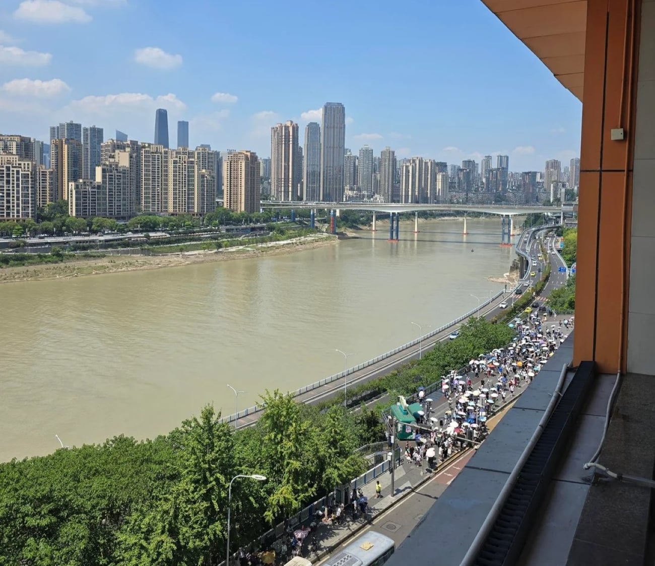A city skyline with tall buildings, a river, and a bridge. Crowds gather along a riverside road with many umbrellas, trees, and vehicles visible under a clear, sunny sky—capturing the vibrant energy often seen in some of the Biggest Cities In The World.