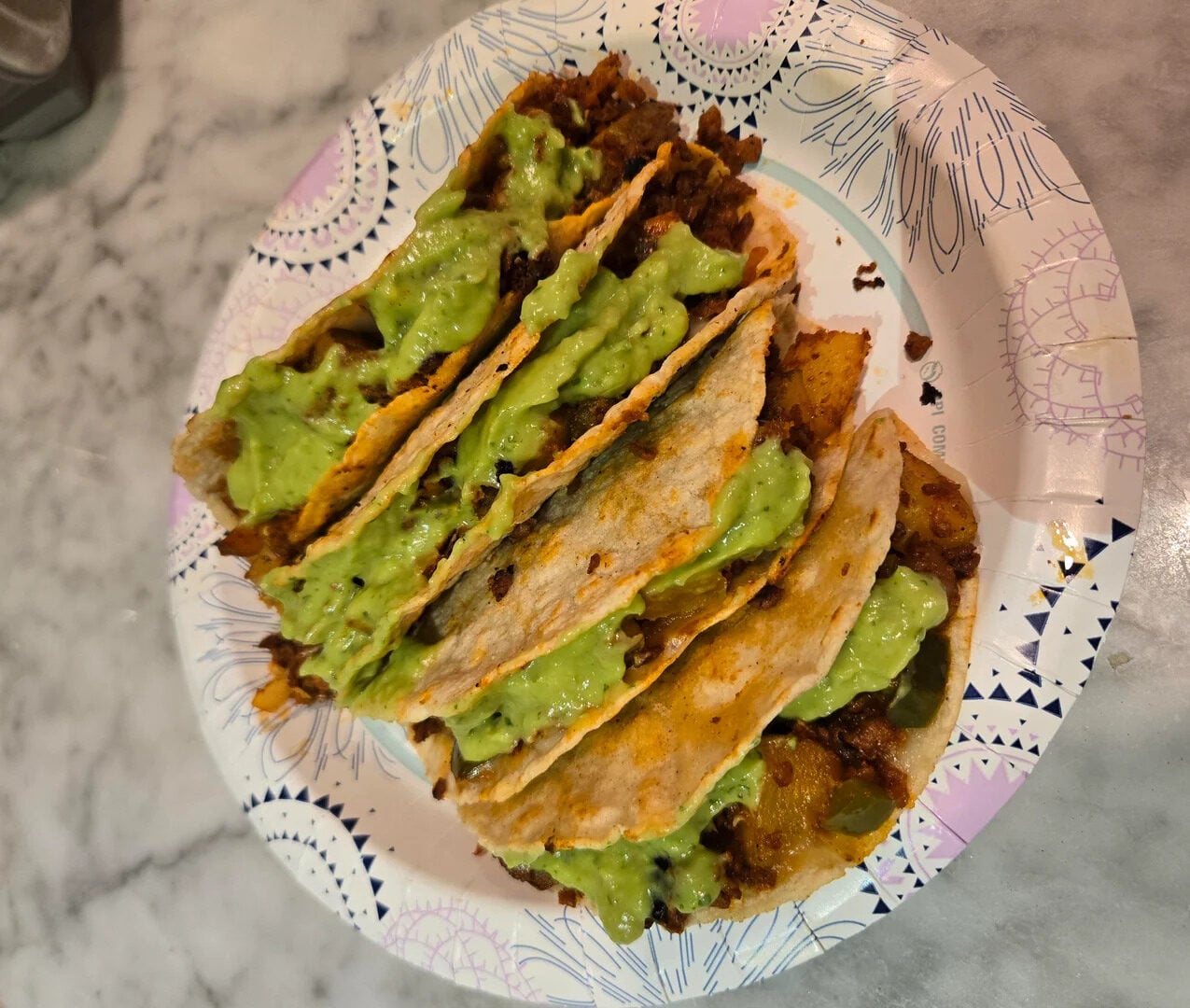 Three tacos filled with seasoned meat and topped with green guacamole, served on a decorative paper plate on a marble countertop.