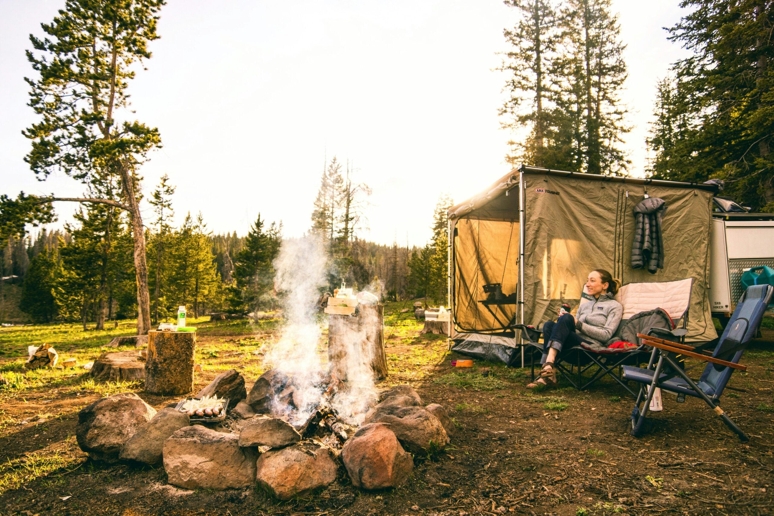A person relaxes in a camping chair by a fire pit, with a tent and tall trees in the background, under a sunny sky. Smoke rises from the fire, and camping items are scattered around the site.
