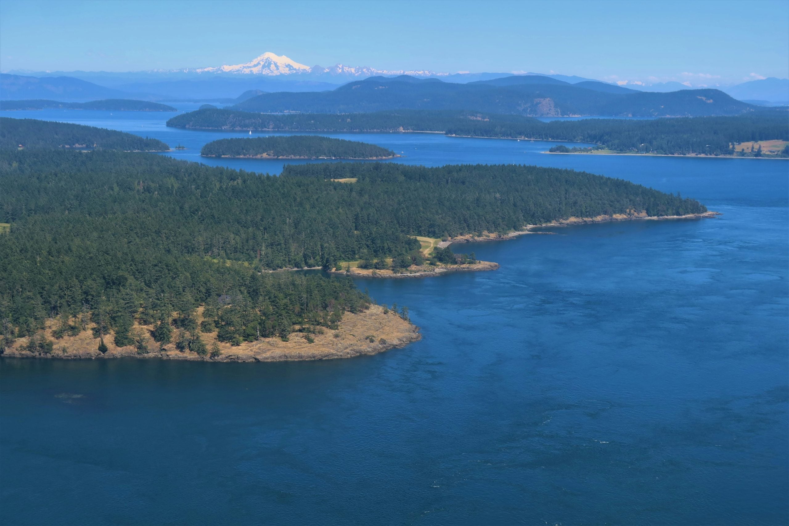 Aerial view of forested islands surrounded by blue water, with distant mountains and a snow-capped peak on the horizon under a clear blue sky.