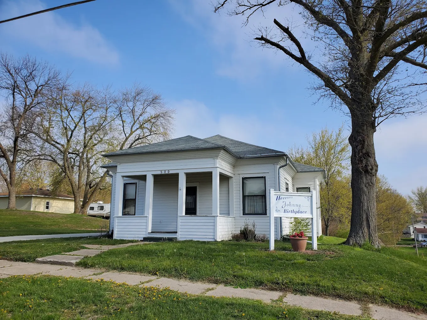 A small white house with a covered porch and sign reading “Museum Johnny Birthplace” sits on a grassy lot with trees and a sidewalk under a blue sky. Affordable small towns for retirement.