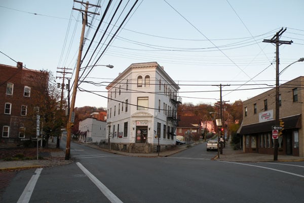A white three-story building stands at a street intersection, surrounded by power lines, brick, and beige buildings. Trees and a hill are visible in the background under a clear sky.