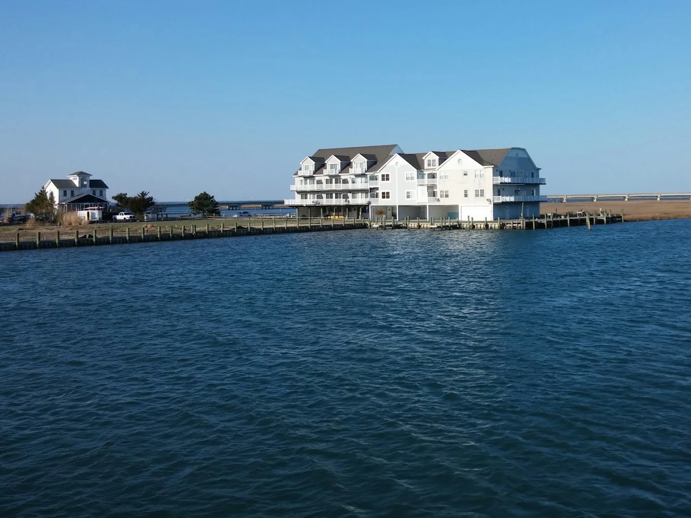 A large white building with balconies sits on the water’s edge, surrounded by a wooden dock. There is another smaller house nearby and a clear blue sky overhead.
