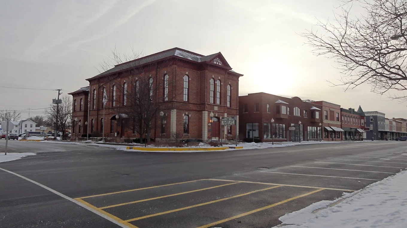 A quiet, snowy small-town street with historic brick buildings, empty parking spaces, and leafless trees on a cloudy day.