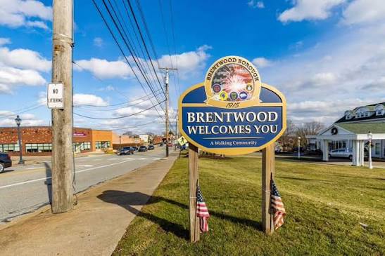 A “Brentwood Welcomes You” sign stands by a sidewalk and road, with two American flags at its base. Buildings and power lines are visible under a partly cloudy sky.