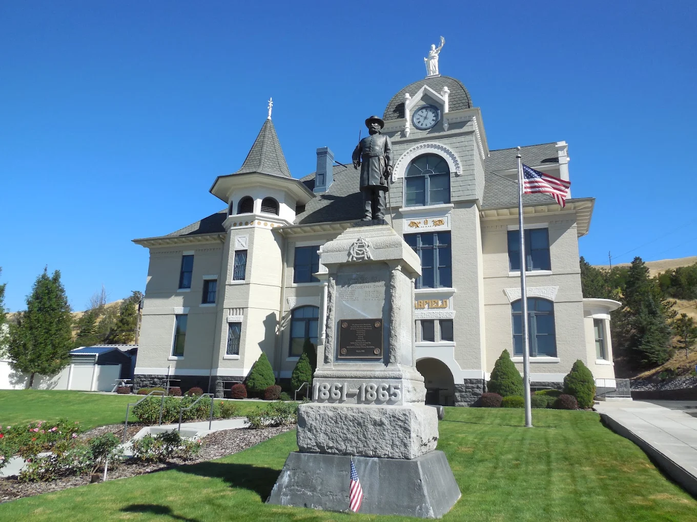 A historic courthouse with a clock tower stands behind a Civil War soldier statue and memorial, with American flags and manicured lawns under a clear blue sky.