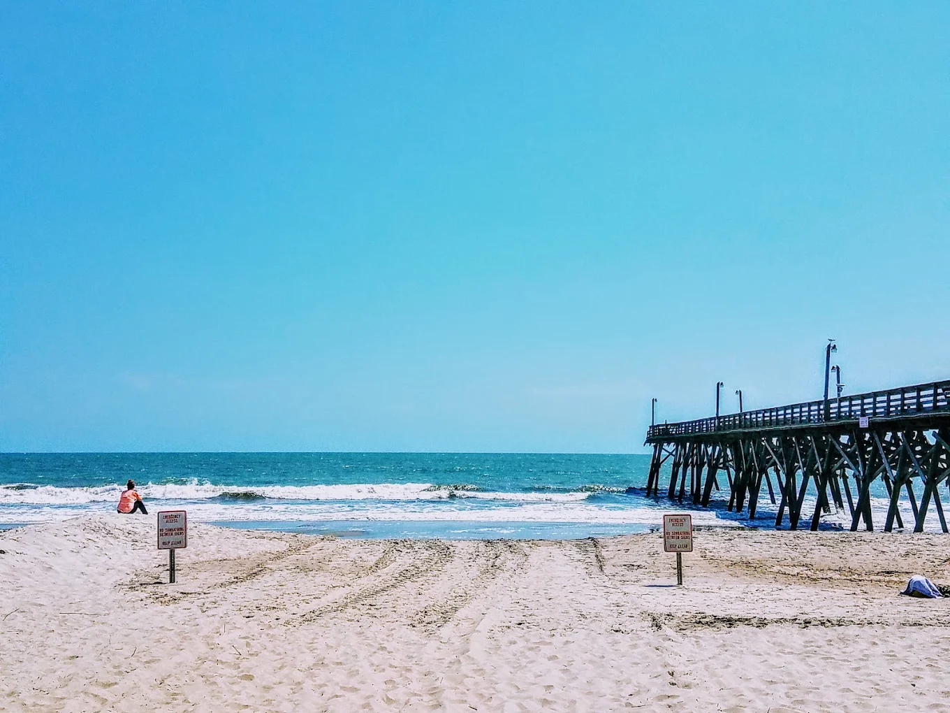 A sandy beach with two warning signs, a person sitting near the shore, waves in the ocean, and a wooden pier extending from the right into the water under a clear blue sky.