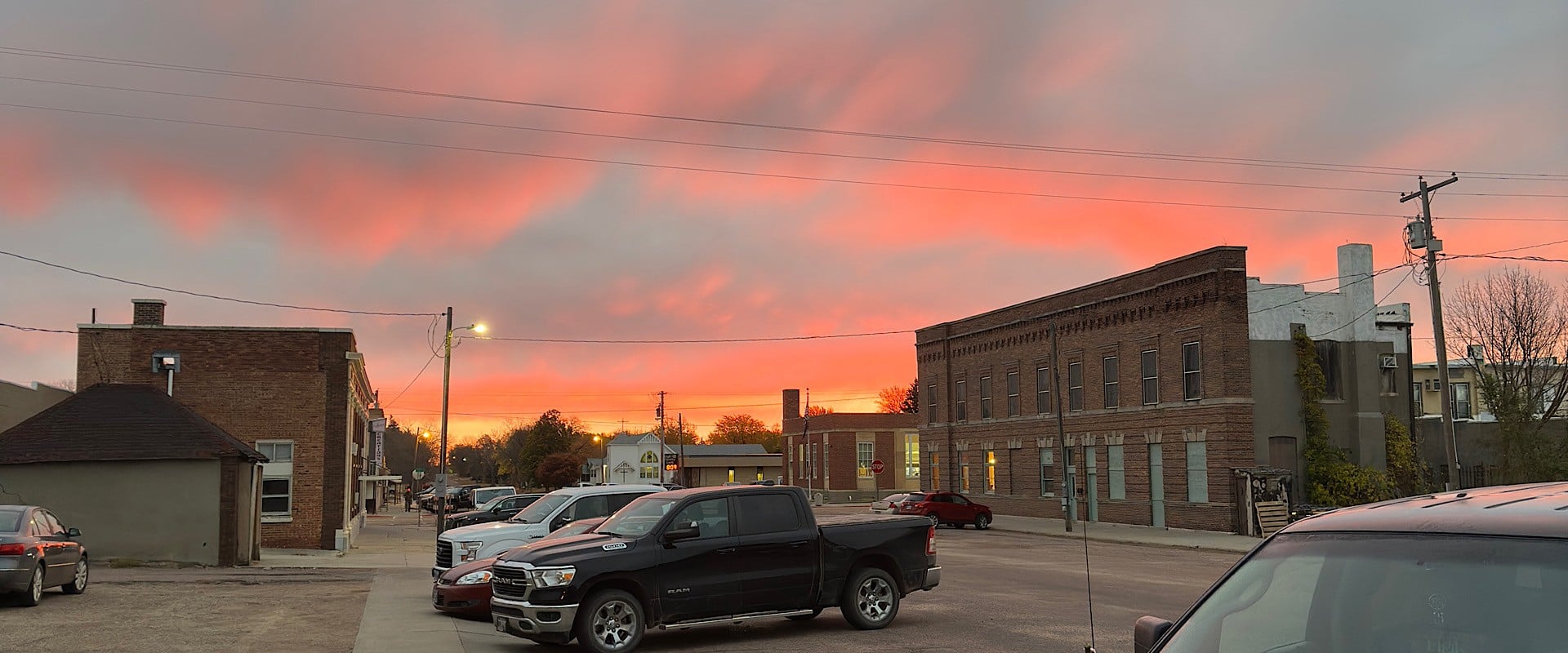 A small town street at sunset, with parked cars and brick buildings lining both sides. The sky is vibrant with orange and pink clouds, casting a warm glow over the scene.