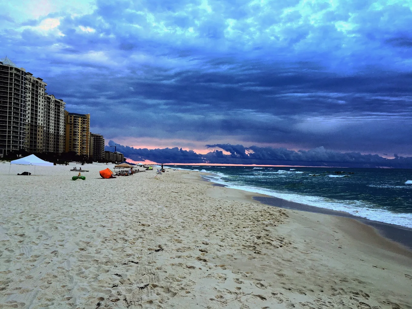 A sandy beach with gentle waves, high-rise buildings on the left, and dramatic clouds above. The sky shows hints of sunset colors near the horizon.