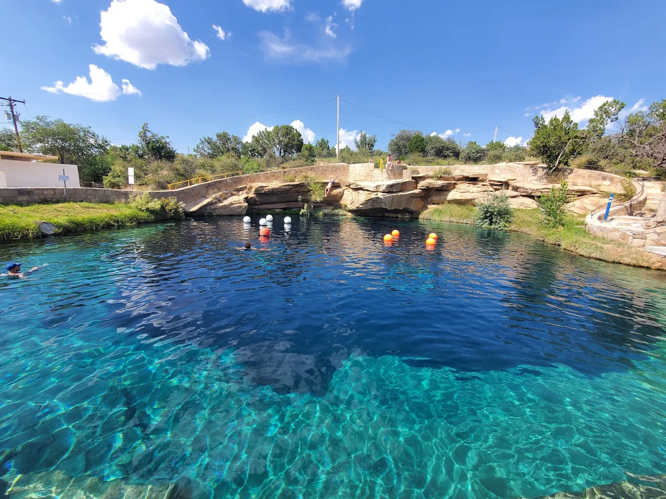 A clear blue spring pool surrounded by rocky edges, green shrubs, and a bright sky. Floating buoys and a swimmer are visible in the water.