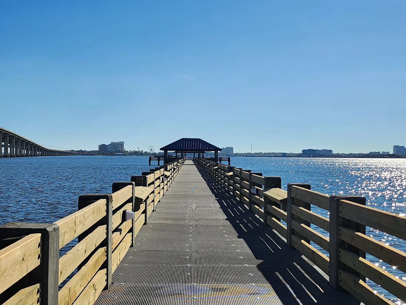 A wooden pier with railings extends over calm blue water toward a covered gazebo, with city buildings visible in the distant background under a clear, bright sky. Affordable beach towns for retirees.