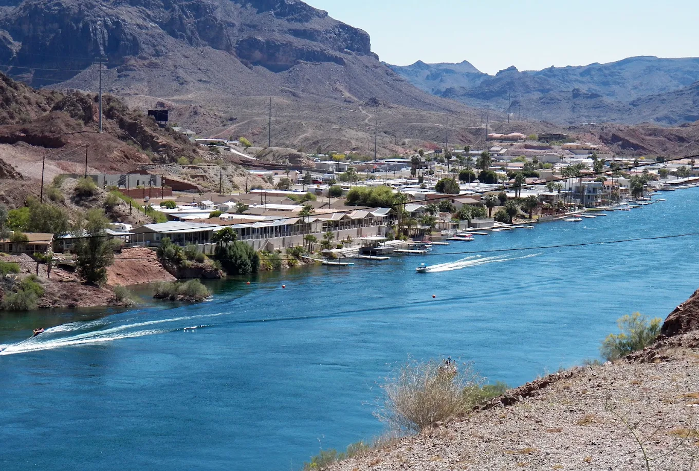 A vibrant blue river with boats runs alongside a small town, with houses and trees nestled between arid, rocky mountains under a clear sky.