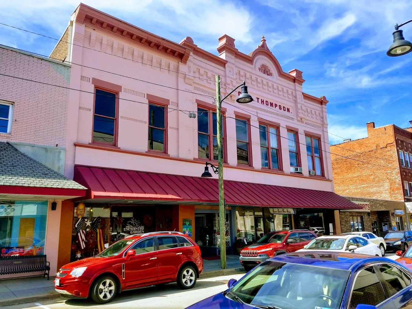 Historic pink building with “Thompson” sign, large arched windows, and red awning. Several cars are parked on the street in front under a bright blue sky.