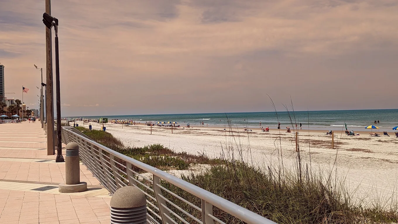A sandy beach with people, blue umbrellas, and tall grasses next to a boardwalk with railings and streetlights under a cloudy sky. The ocean is calm and stretches to the horizon.