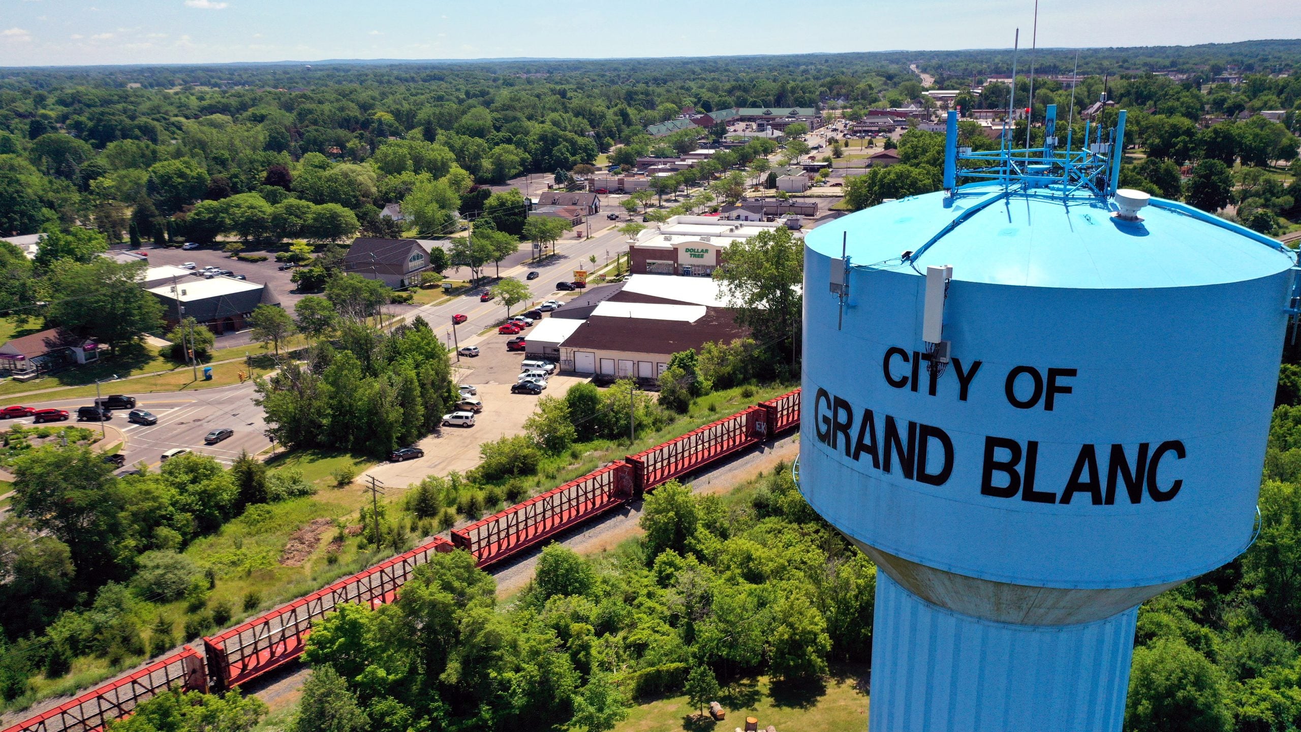 A large blue water tower labeled “City of Grand Blanc” stands above green trees, with a red train passing by below and a town visible in the background under a clear sky.