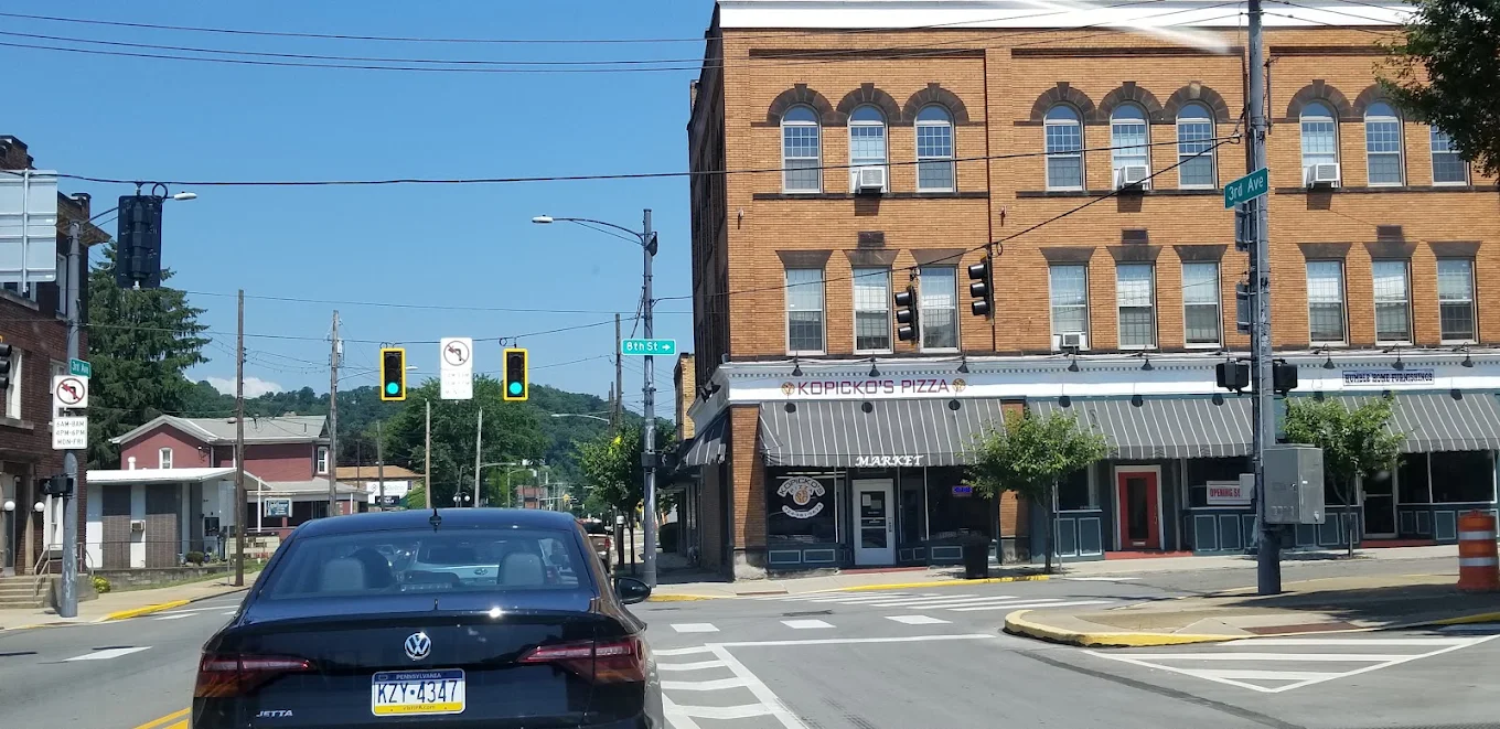 A black car waits at a red light on a sunny day near a brick building with a “Korbecki’s Pizza” sign. Green hills are visible in the background.
