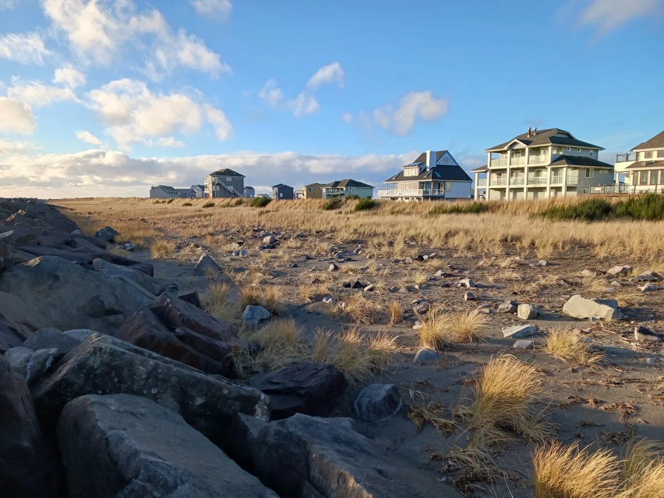 Beachfront houses with large windows sit behind grassy dunes under a blue sky with scattered clouds. Large rocks and dry grass cover the sandy ground in the foreground. The scene is sunny and peaceful.