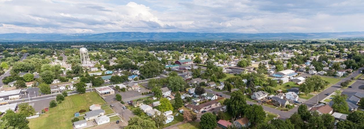 Aerial view of a small town with scattered houses, trees, a water tower, and open fields, with hills and a cloudy sky in the distance.