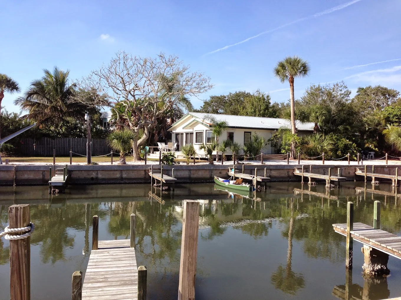 A small house with palm trees sits across a calm canal with wooden docks. A boat with two people is on the water. The sky is clear and blue.