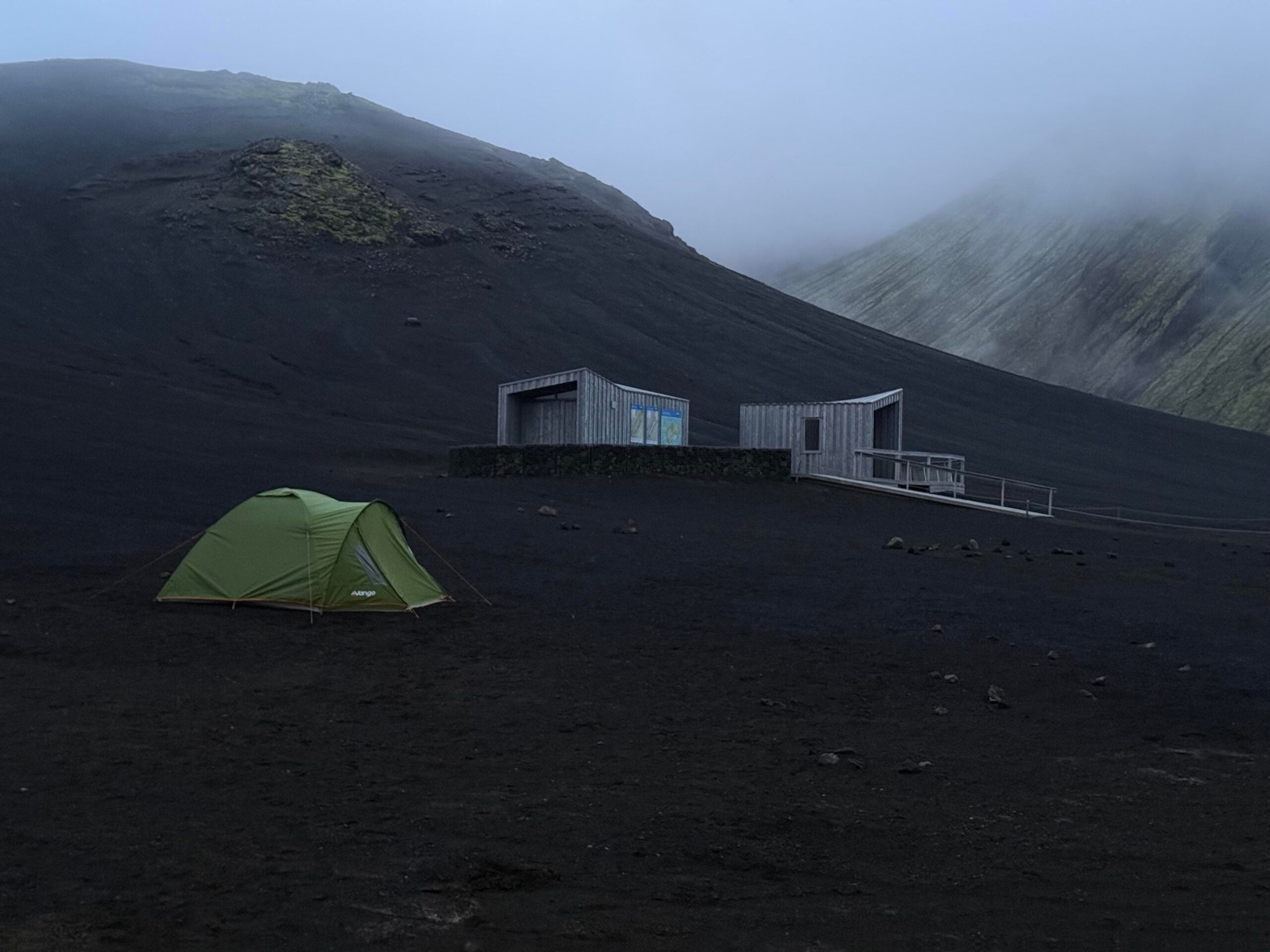 A green tent sits on dark volcanic soil near two small wooden buildings with mountains and mist in the background.