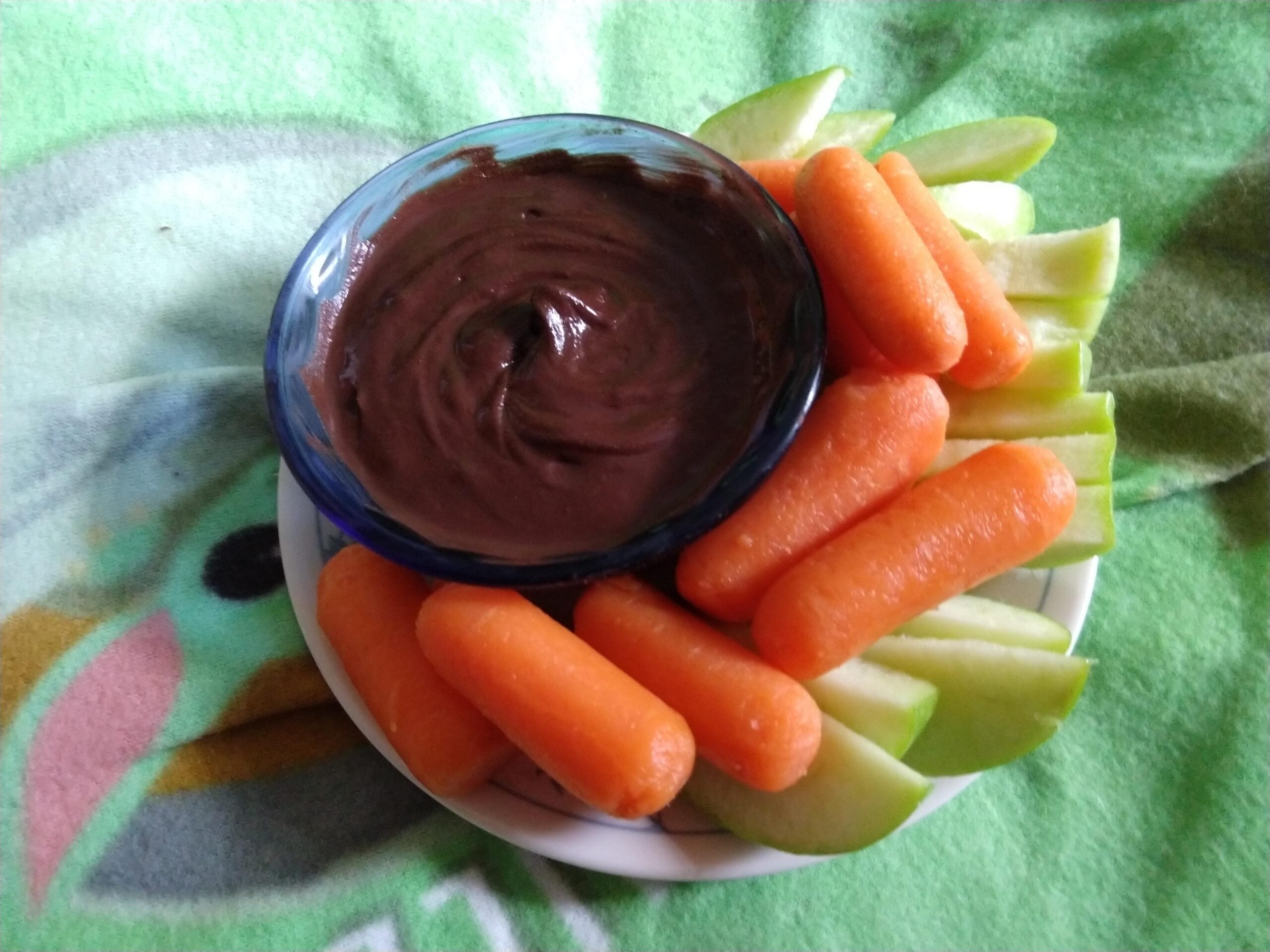 A small plate with baby carrots and green apple slices, arranged around a bowl of chocolate dip, sits on a colorful, patterned surface.