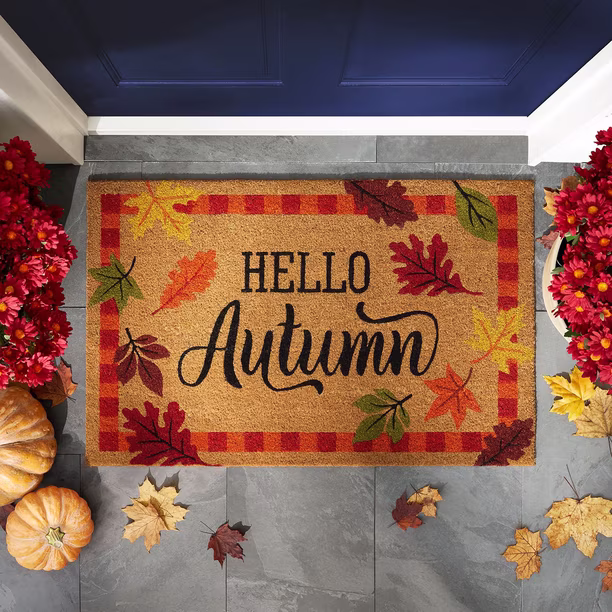 A doormat with “Hello Autumn” in cursive, surrounded by colorful leaves and a red plaid border, placed by a blue door. Pumpkins and red flowers are on each side. Autumn leaves are scattered on the gray stone floor. Sam’s Club fall home finds.