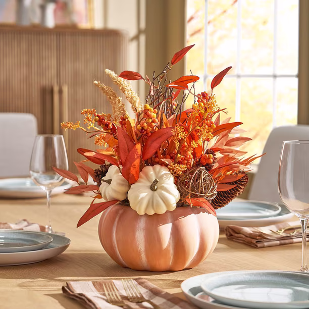 A pumpkin-shaped vase filled with autumn leaves, white pumpkins, and berries sits as a centerpiece on a dining table set with plates, glasses, and napkins.