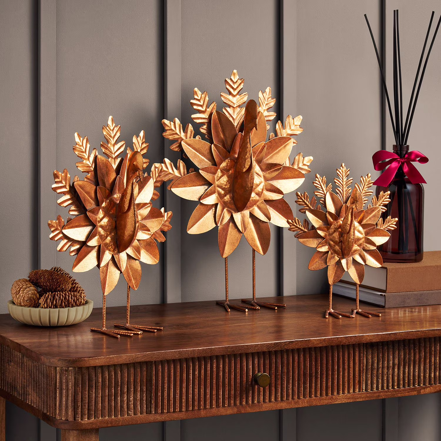 Three gold metal turkey sculptures of varying sizes are displayed on a wooden console table, alongside a bowl of pinecones and a reed diffuser with a red bow, against a gray paneled wall.